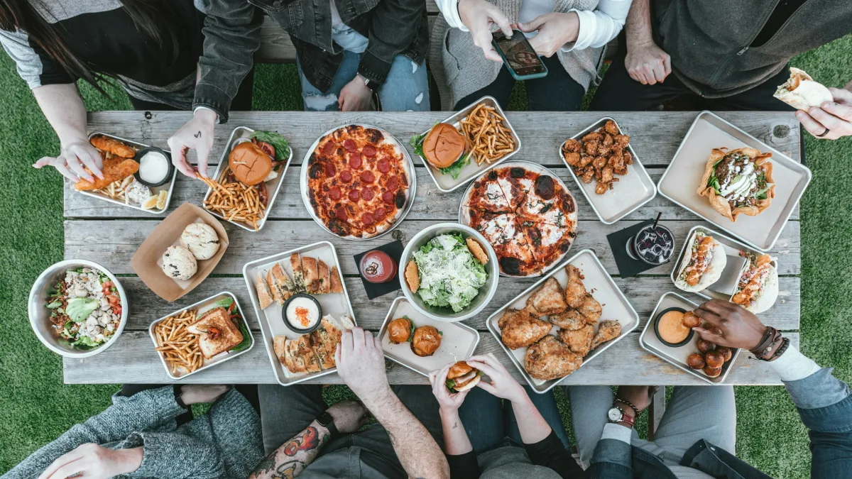 Overhead view of a diverse group sharing pizza, burgers, fries, chicken wings, and salad at an outdoor picnic table.