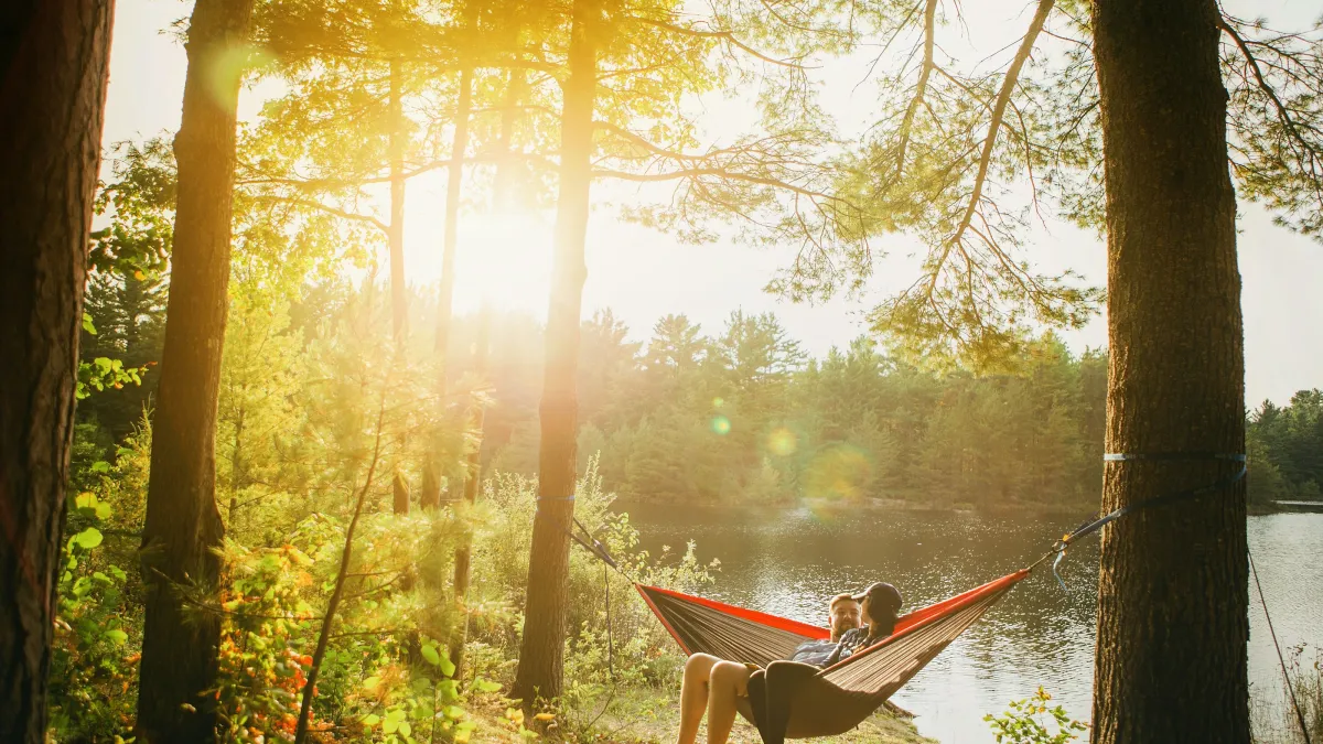 Couple relaxing in a hammock between trees by a sunlit lake with green forest in the background.