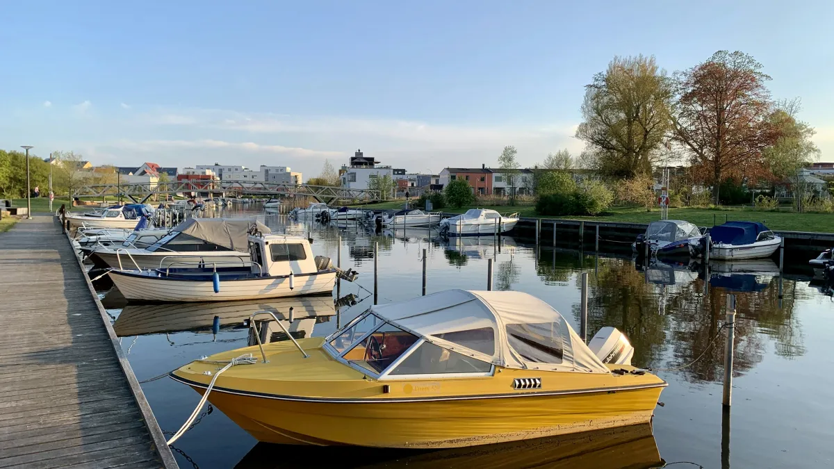Yellow motorboat docked at a marina with other boats and calm water reflecting trees and buildings.