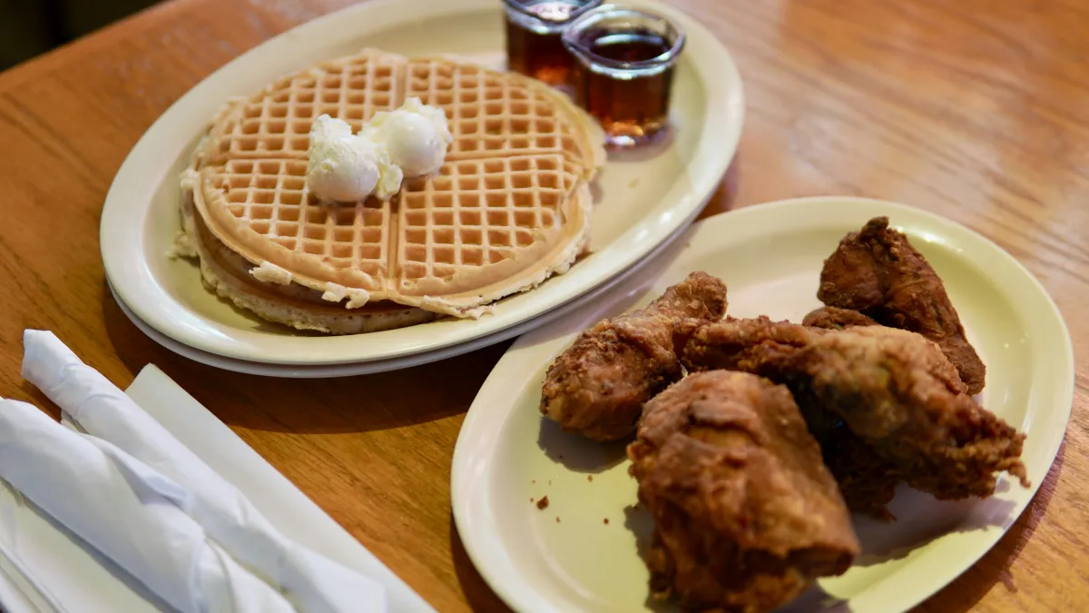 Two plates on wooden table with waffles topped with butter and syrup, and crispy fried chicken pieces.