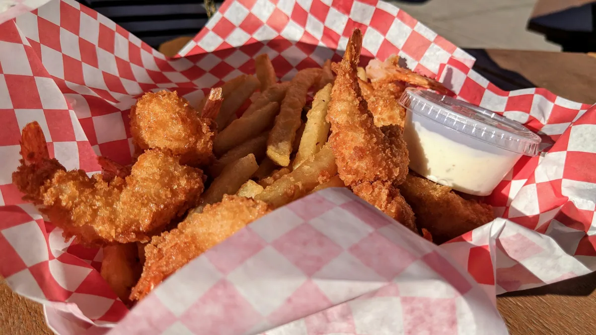 Basket of crispy fried shrimp, French fries, and a container of dipping sauce on red and white checkered paper.
