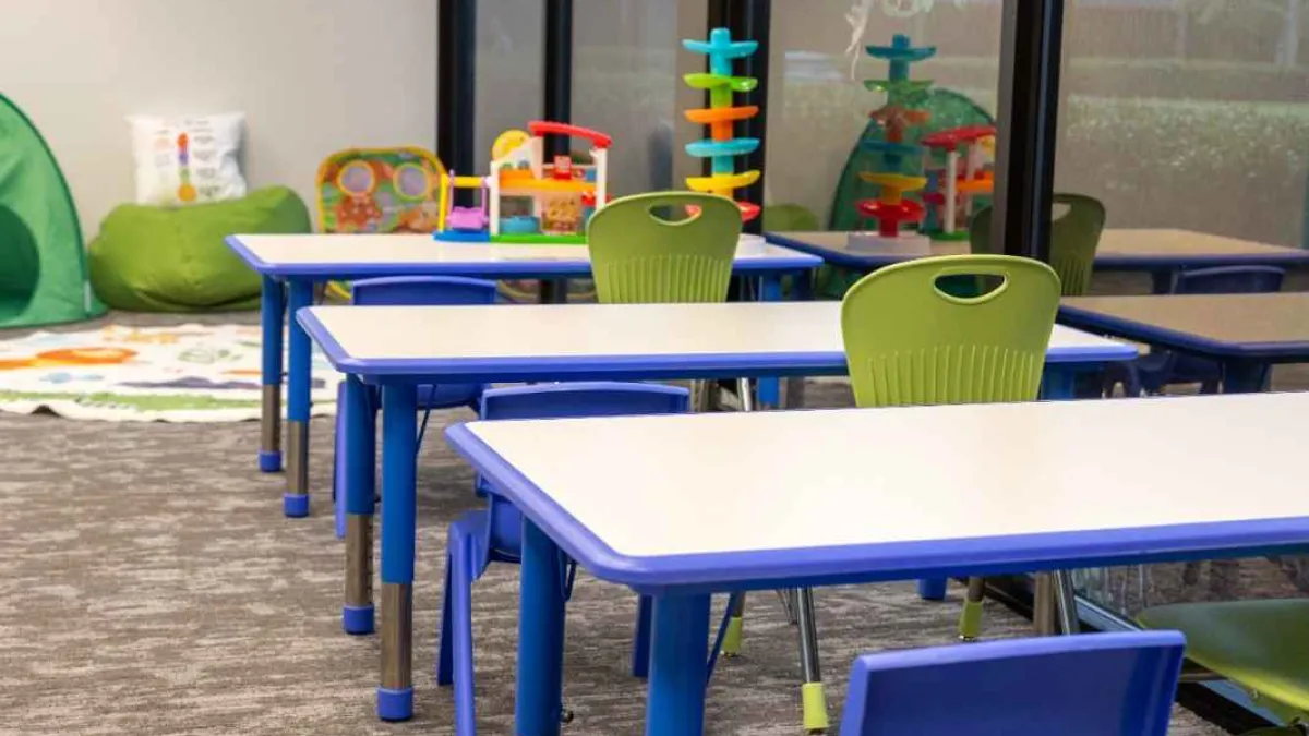 Bright classroom with blue and green chairs, white tables, and children's toys near large windows.
