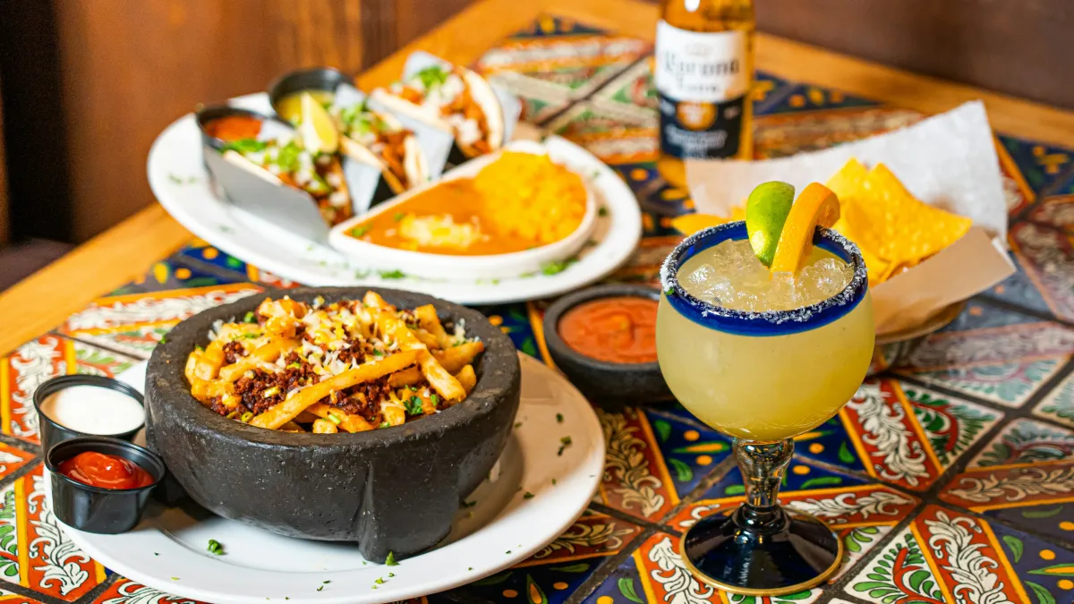 Table with loaded fries in a stone bowl, tacos with rice, margarita, chips, salsa, and a Corona beer on colorful tiled surface.