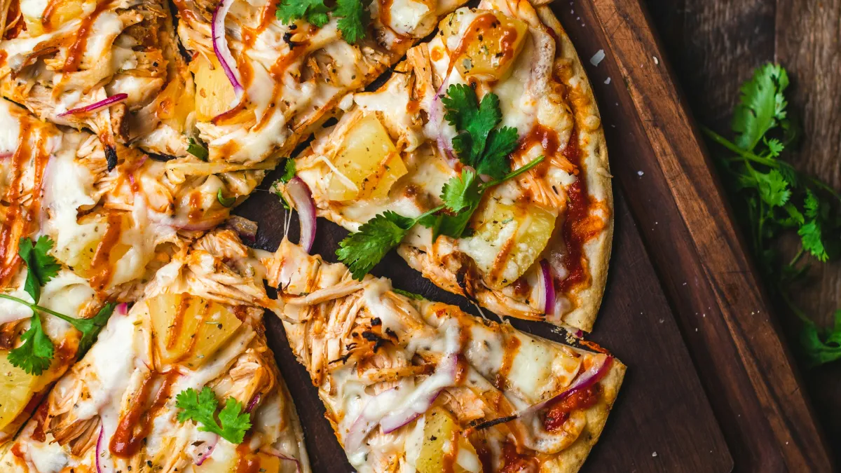 Sliced barbecue chicken pizza with pineapple, red onion, and cilantro on wooden board with garlic and purple onion.