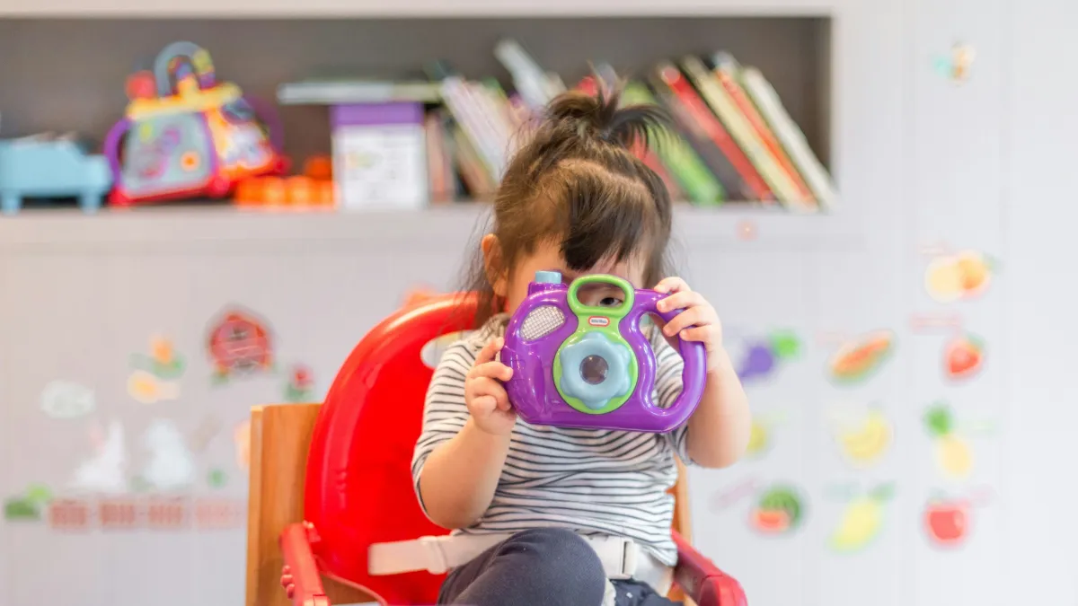 Toddler sitting in red chair holding and looking through a purple and green toy camera indoors.