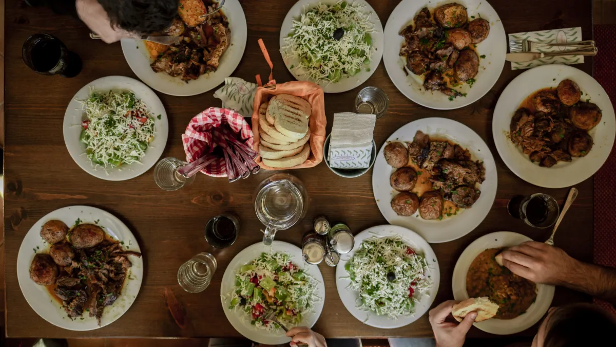 Overhead view of a wooden table with plates of salad, meat, round potatoes, bread, and drinks being shared by diners.