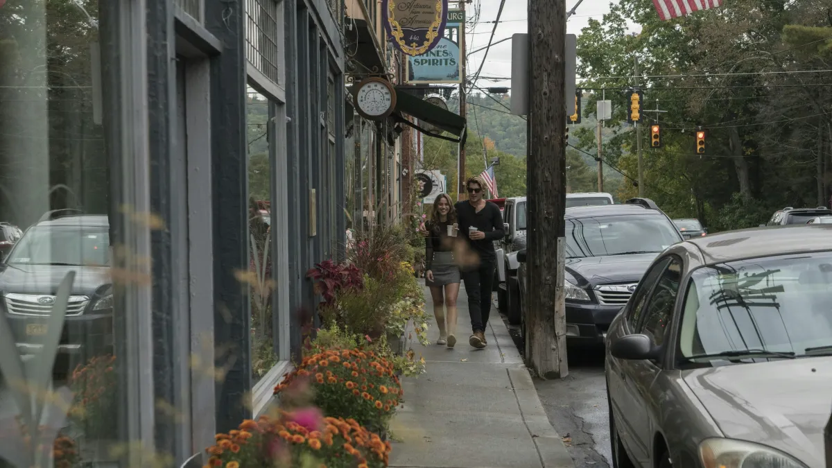 Couple walking on a small town sidewalk with flowers and parked cars on a cloudy day.