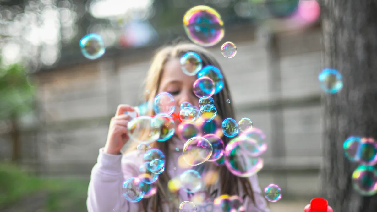 Young girl blowing colorful soap bubbles outdoors in a backyard with wooden fence background.