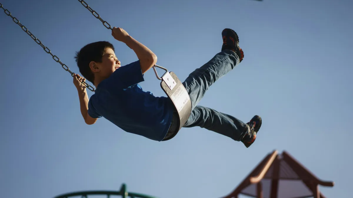 Boy in blue shirt swinging high on a playground swing against a clear blue sky at sunset.