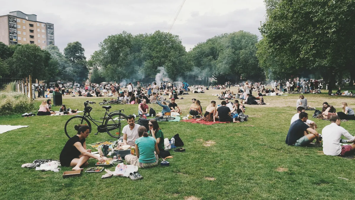 Large crowd of people enjoying a sunny day picnic in a green urban park with trees and buildings nearby.
