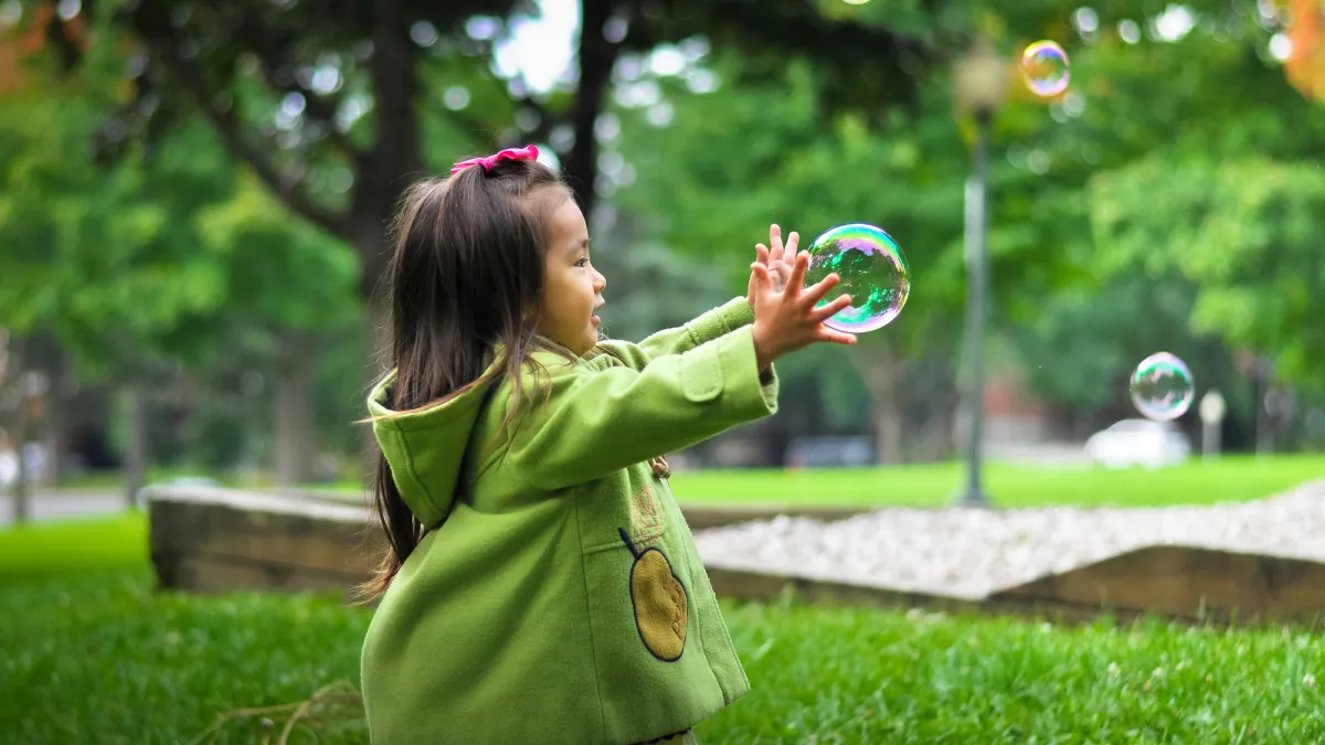 Little girl in green coat reaching out to touch soap bubbles in a park on a bright day