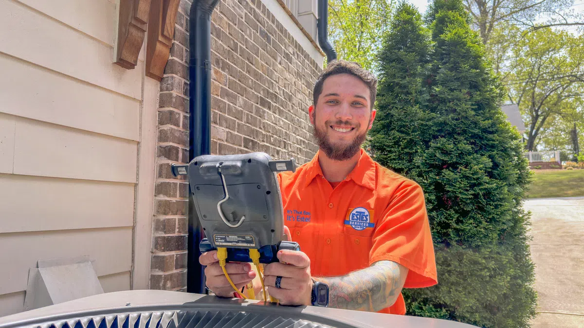 Technician in orange shirt working on HVAC unit outside a residential home on a sunny day.