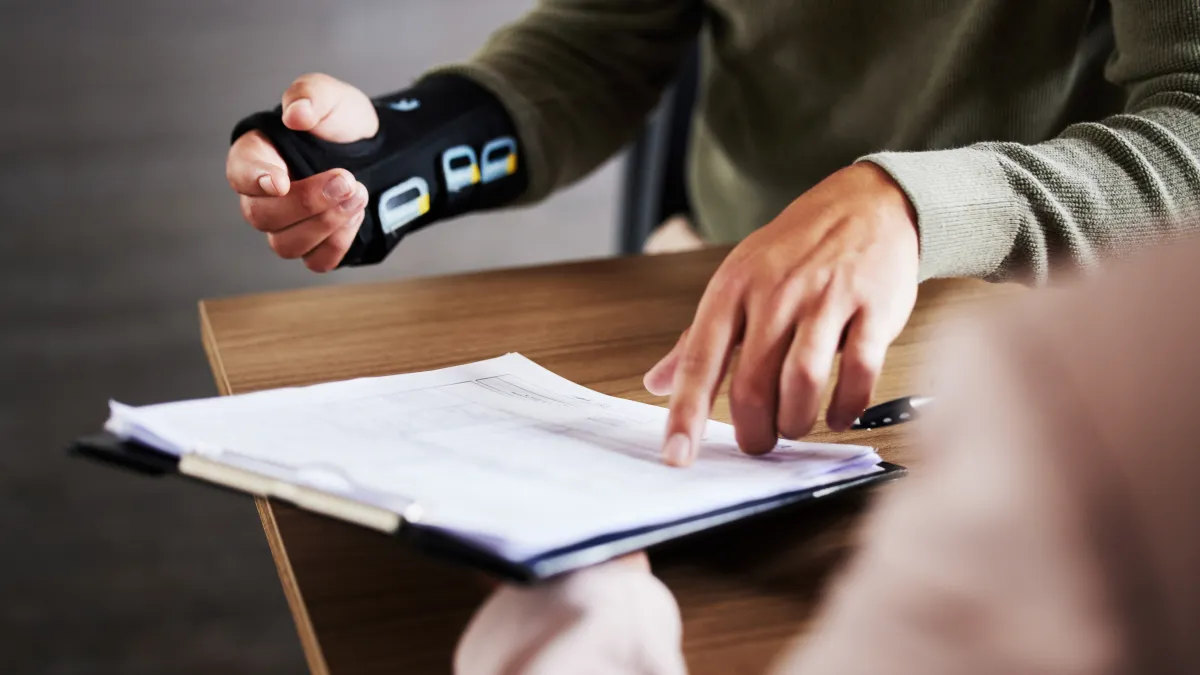 Person wearing wrist brace pointing at document on clipboard during consultation at wooden table