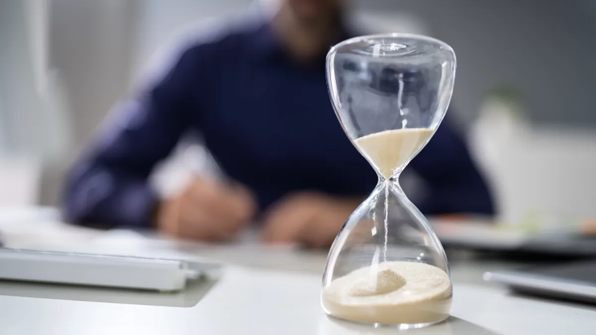 Clear hourglass with sand running through on desk, blurred person writing in background, business setting.