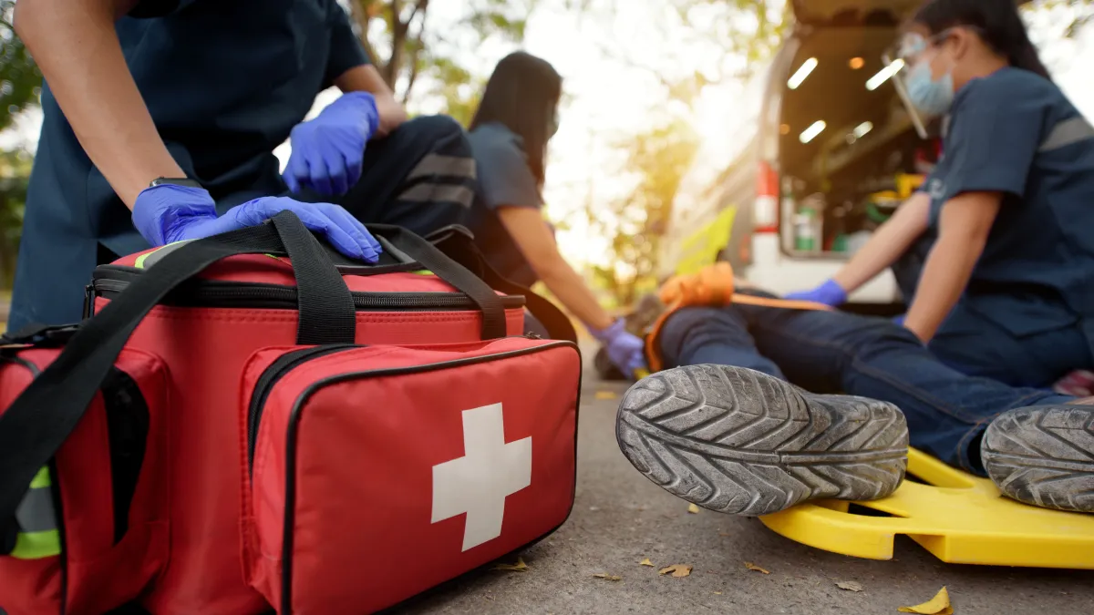 Emergency responders wearing gloves and masks attending to injured person on stretcher with first aid kit nearby.