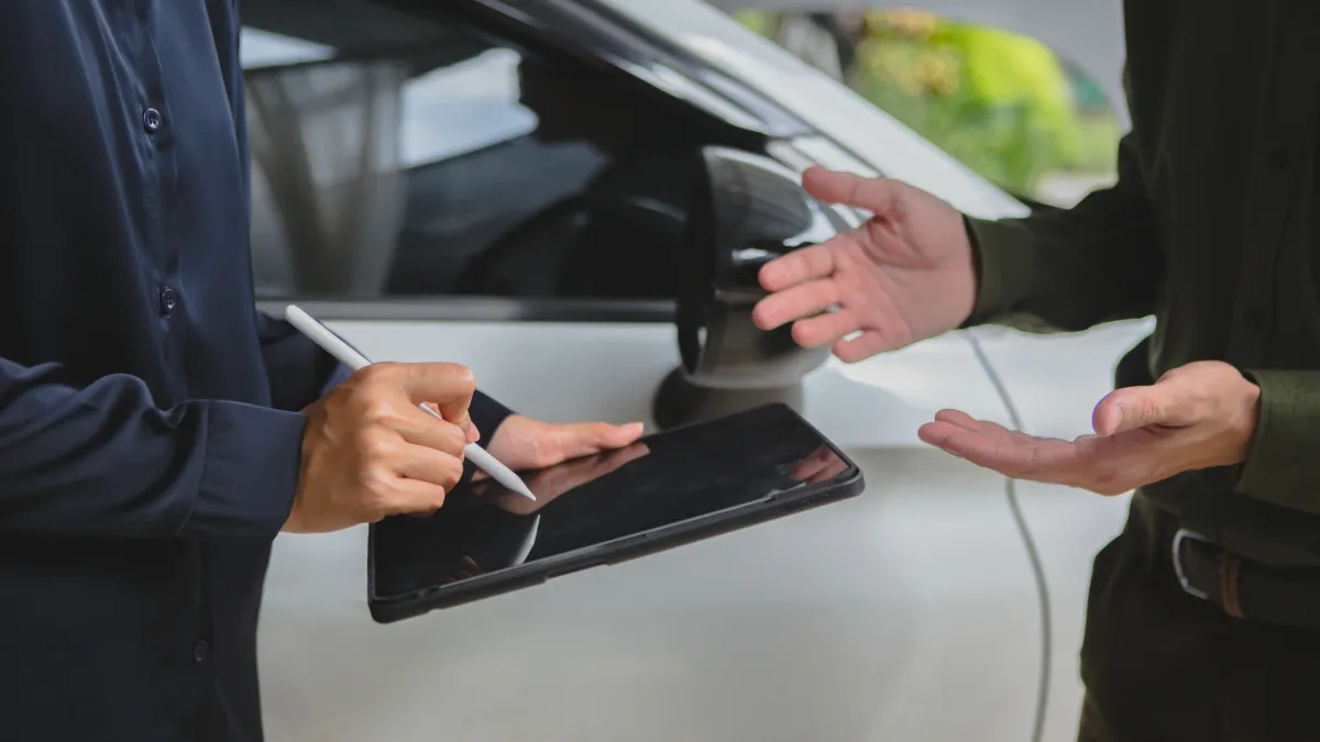 Two people near a car, one signing on a tablet with a stylus while the other gestures with hands