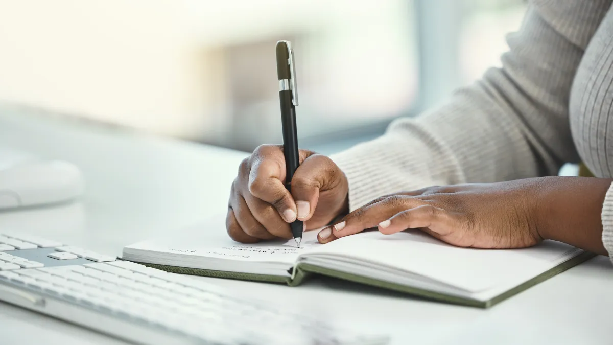 Close-up of a person writing in a notebook with a pen beside a computer keyboard on a white desk.