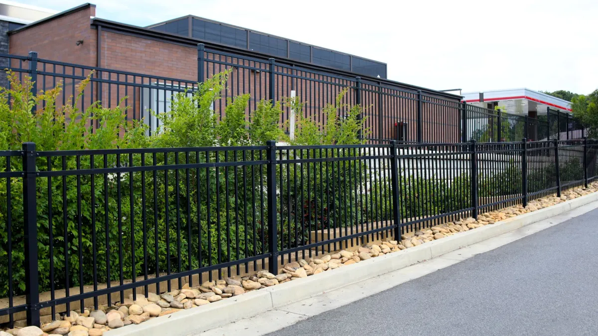 Black metal fence with green shrubs and rocks lining a sidewalk next to a brick building under a clear sky