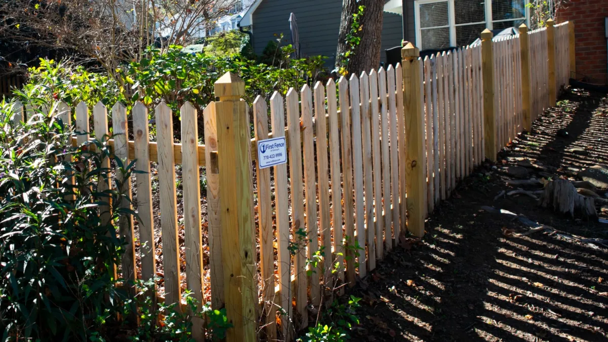 A wooden picket fence bordering a garden with sunlight casting shadows on the ground.