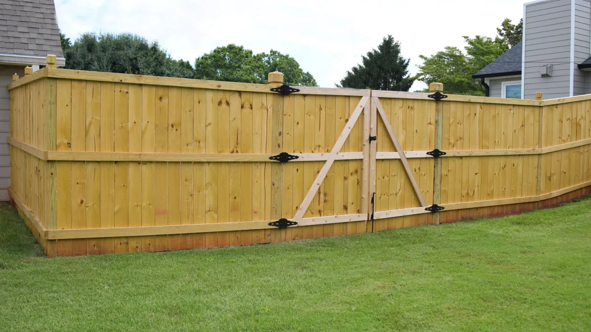 New wooden privacy fence with double gate installed around a backyard lawn area between houses.