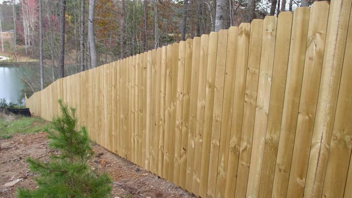 A wooden privacy fence with pointed tops extends along a wooded area beside a tranquil pond.