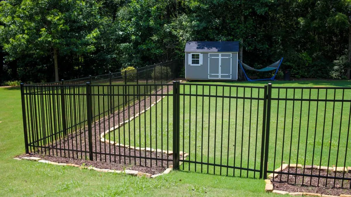 Black metal fence enclosing a green yard with a gray shed and hammock in a backyard with trees.