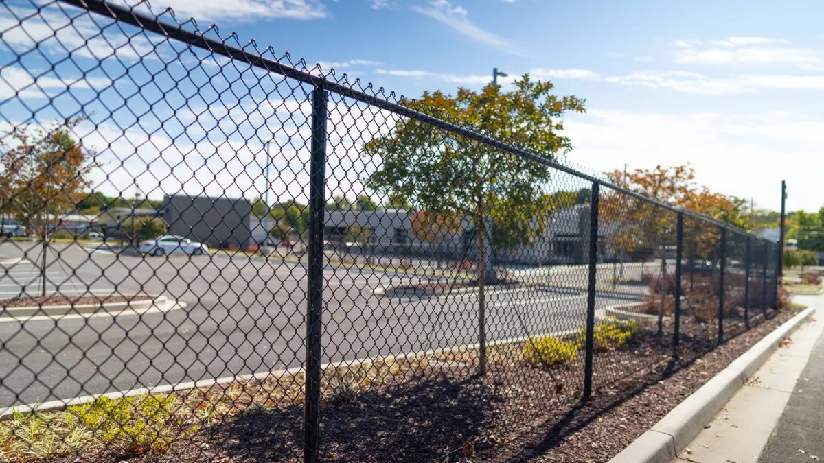 Chain-link fence along a smooth pavement near a parking lot with trees and clear blue sky.