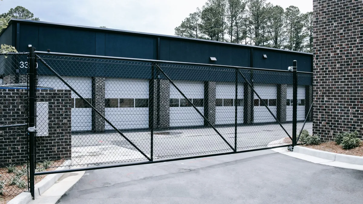 Black chain link fence gate in front of brick building with white garage doors and trees in background.