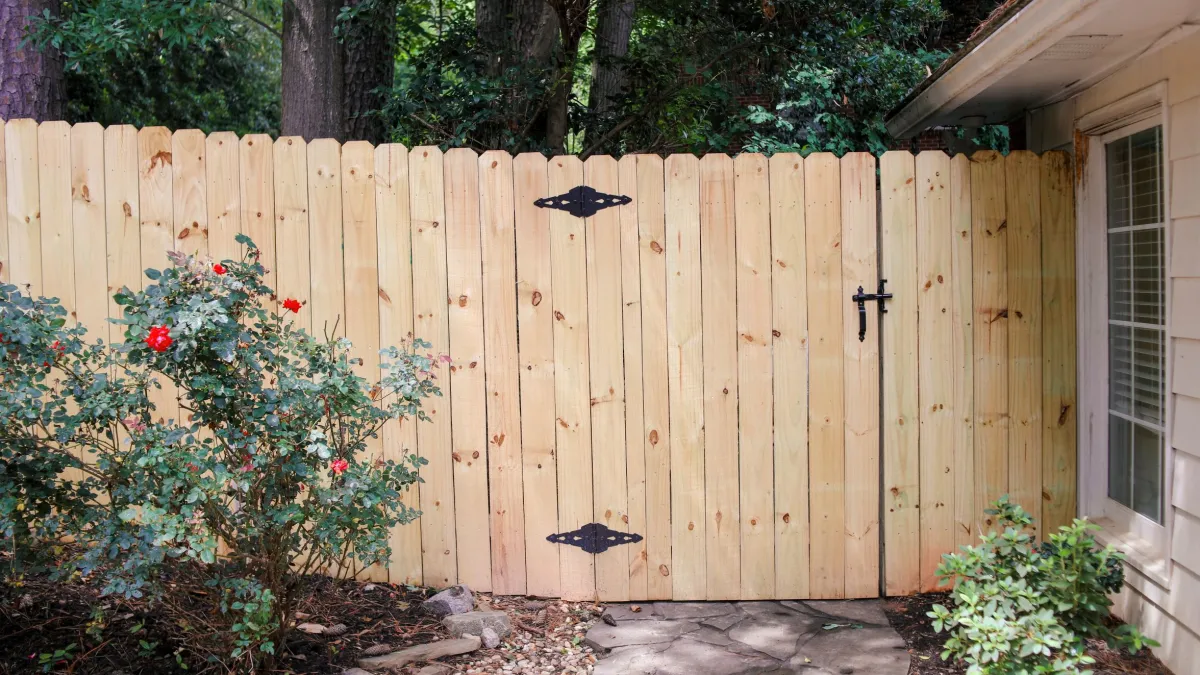 Wooden fence gate with black hinges beside a house, surrounded by bushes and trees in a backyard.