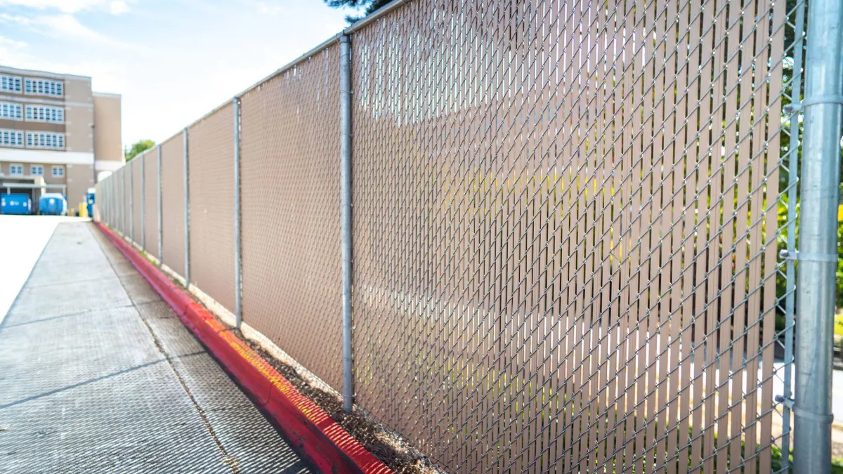A chain-link fence stretches along a walkway beside a school building under a clear blue sky.