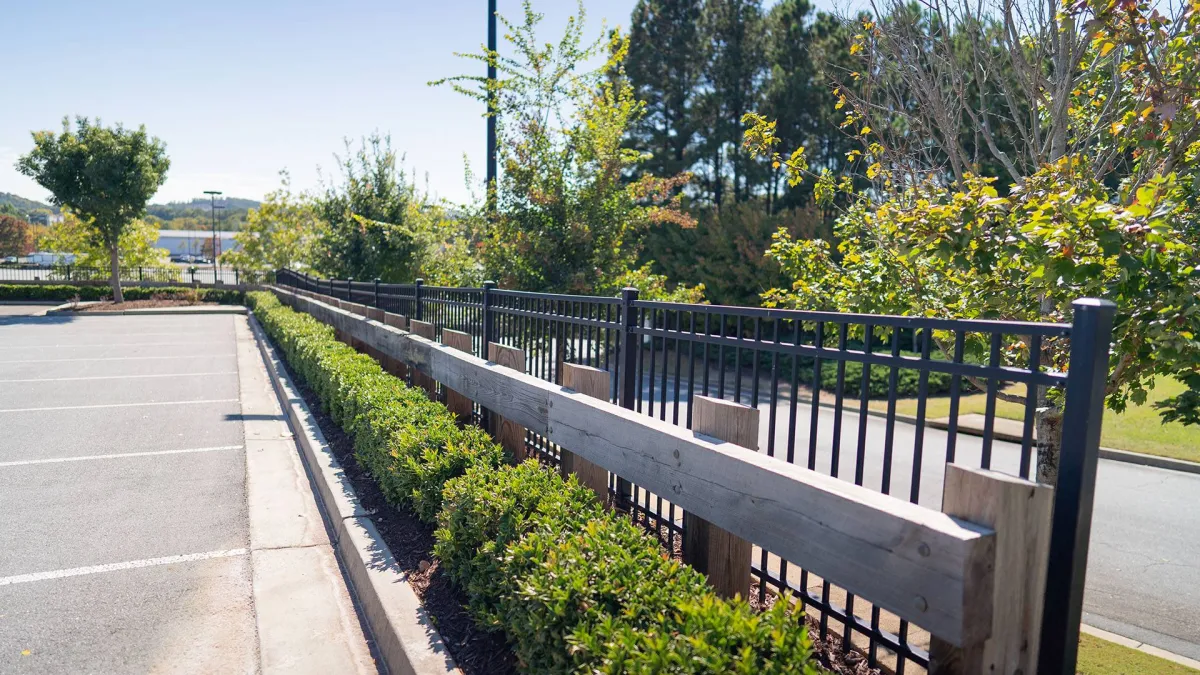 Iron fence along a landscaped area with lush greenery in a sunny outdoor parking lot setting.