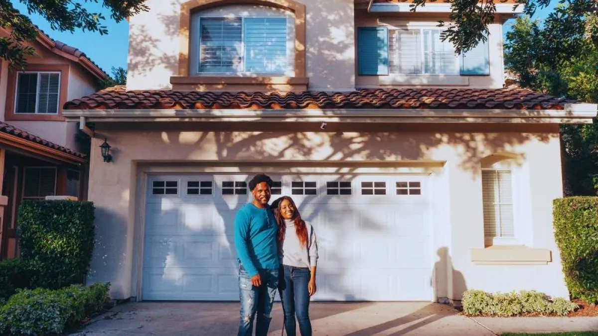 Smiling couple standing in front of a two-story house with a double garage and tile roof on a sunny day.