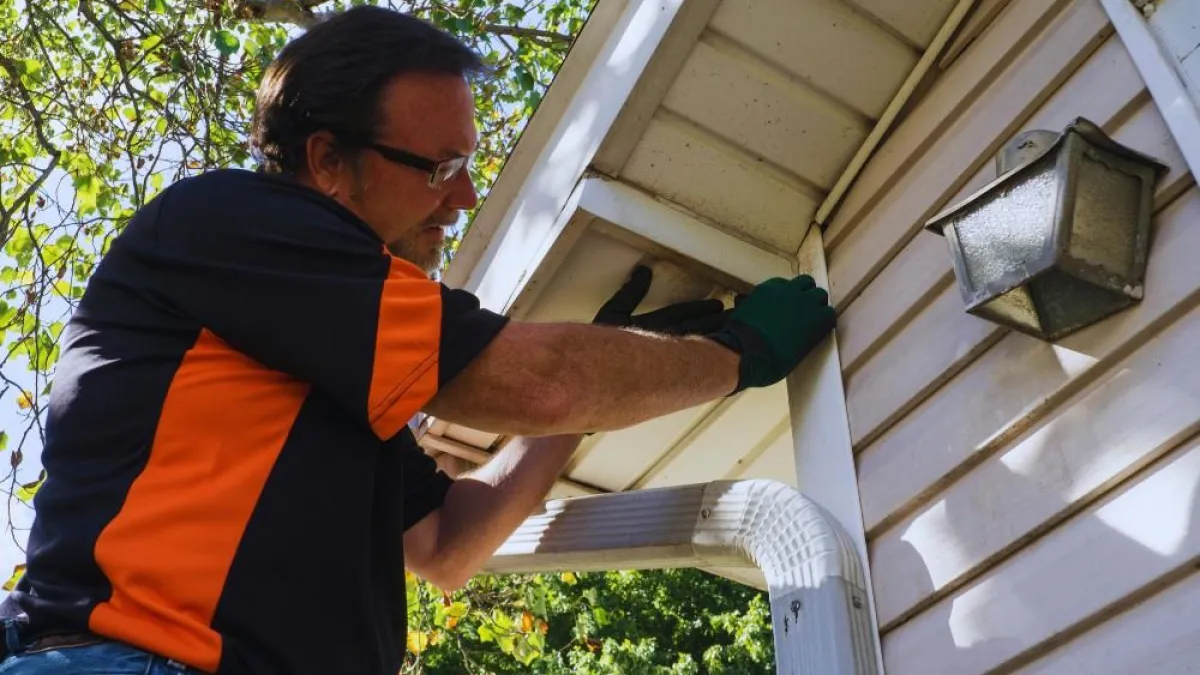 Man wearing gloves inspecting or repairing a house gutter on a sunny day with trees in the background