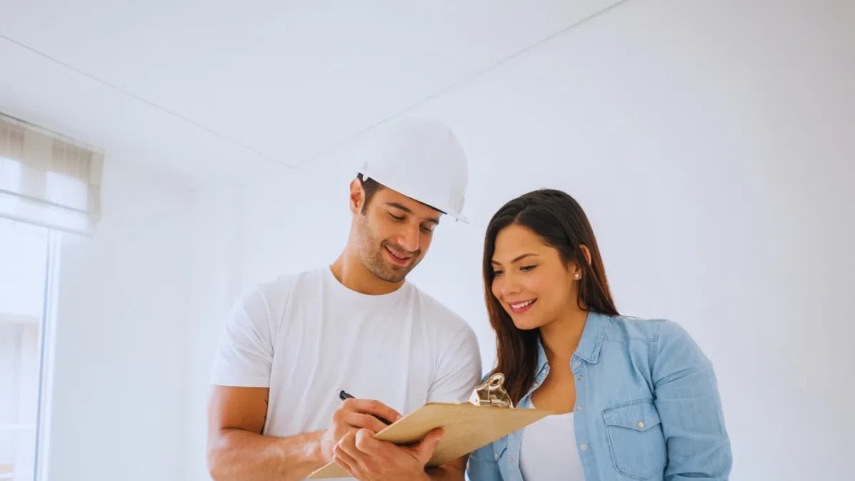 Male contractor in hard hat explaining plans on clipboard to smiling woman in casual clothes indoors.