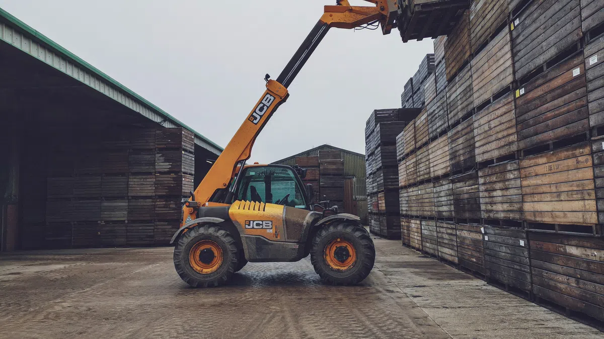 Yellow JCB forklift stacking wooden crates in an industrial warehouse yard on a cloudy day