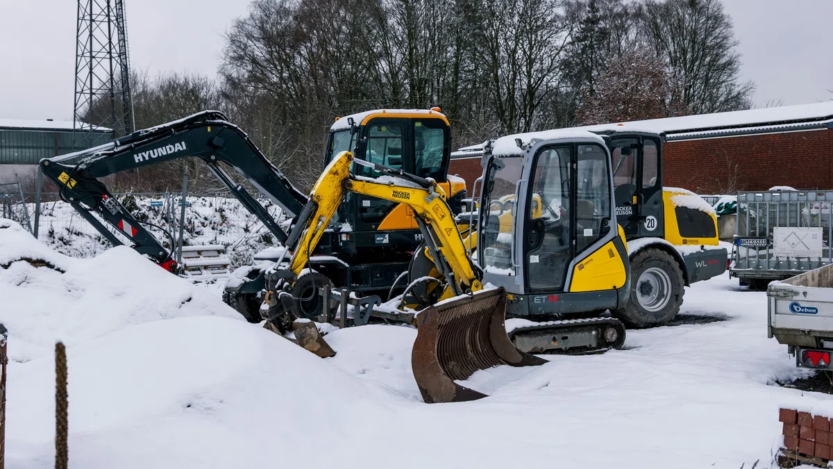 Yellow and black excavators parked on snow-covered ground near construction materials and bare trees in winter.