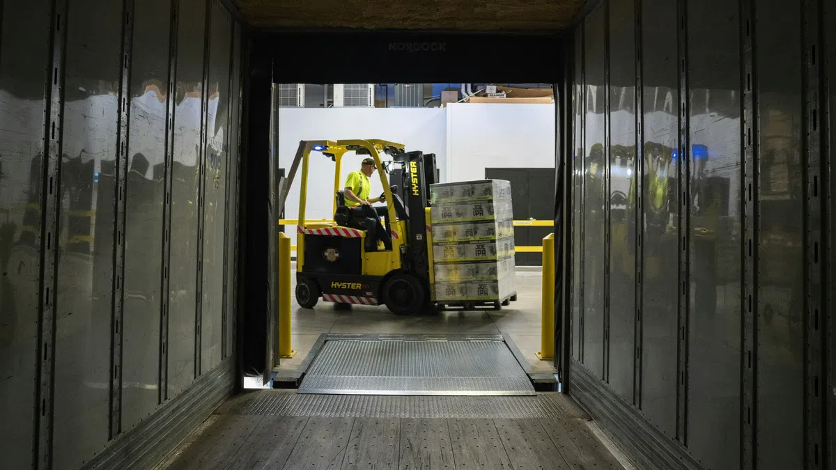 Forklift operator loading stacked pallets into a trailer through a warehouse loading dock with metal walls.