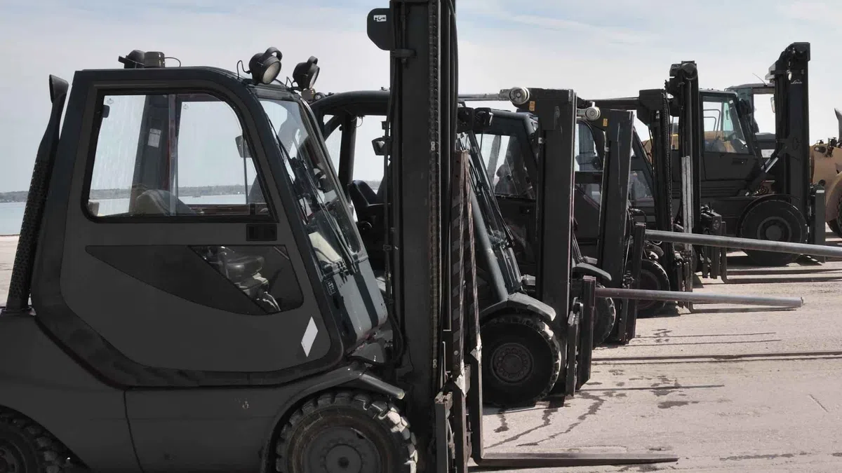 Row of black forklifts parked in a line outdoors on concrete under a cloudy sky.