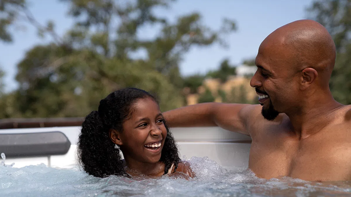 Father and daughter laughing together in an outdoor hot tub on a sunny day with trees in background