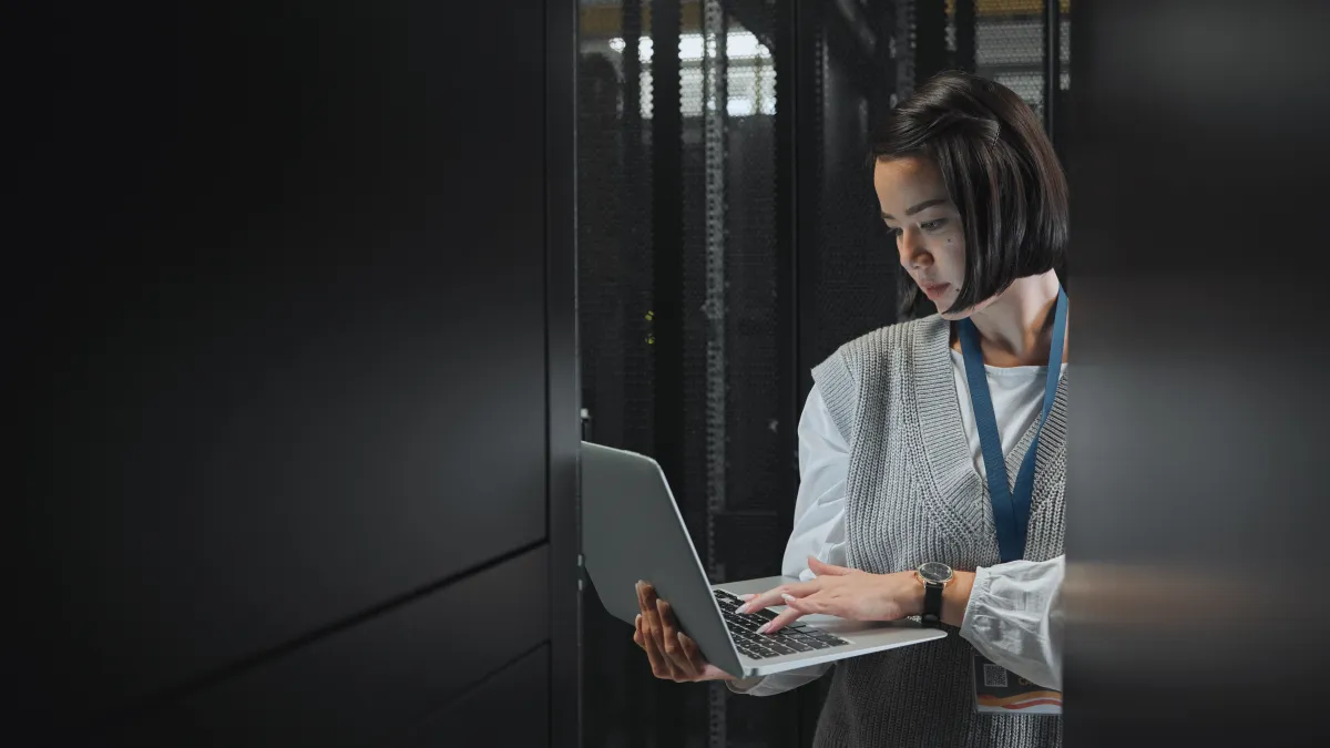 Woman working on laptop inside a server room with data racks in the background.