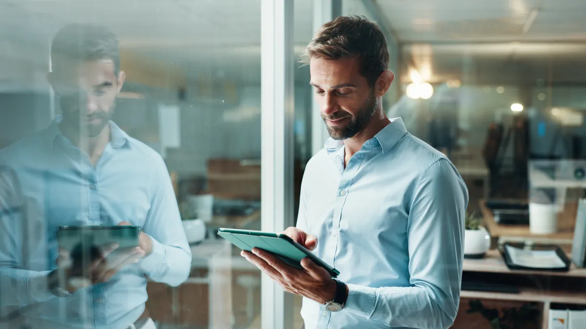 Man in light blue shirt using a digital tablet, reflected in office glass wall with modern workspace background