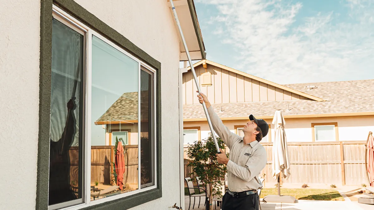 Man cleaning house gutters with a long pole brush on a sunny day beside a white home with succulents.
