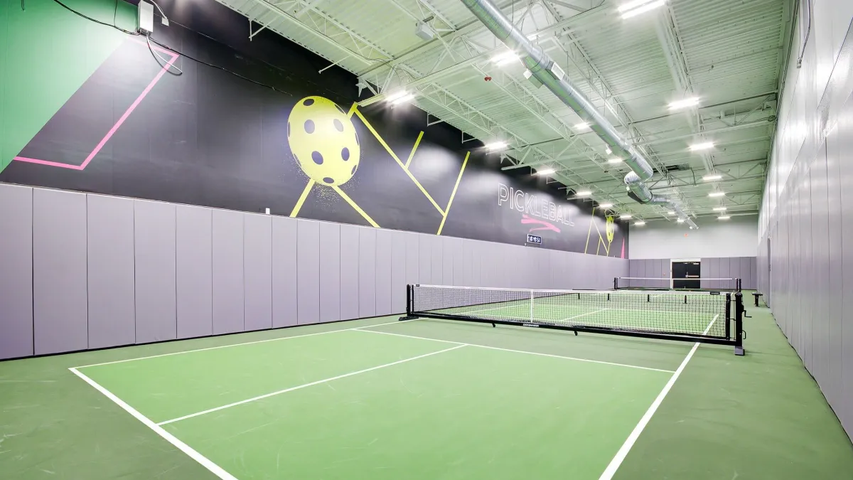 Indoor pickleball court with green flooring, purple wall padding, and bright ceiling lights