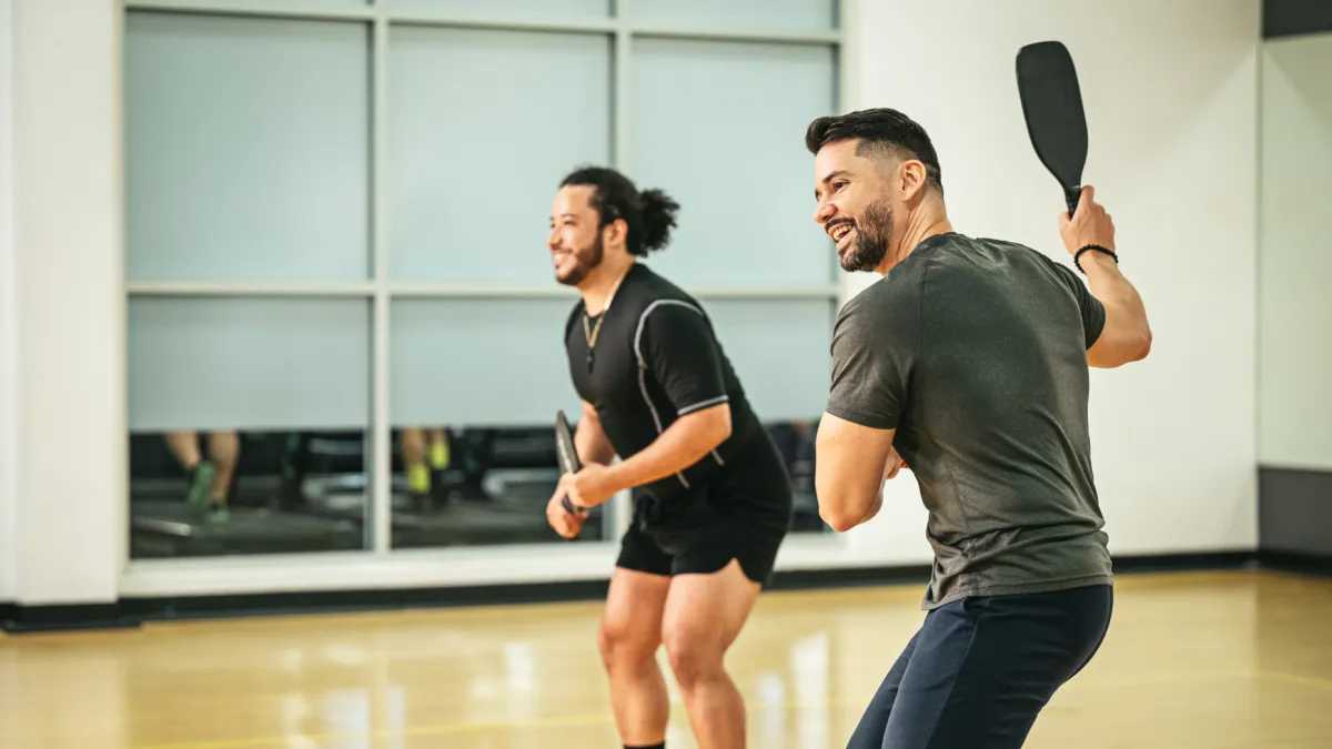 Two men playing pickleball indoors wearing athletic clothing and holding paddles on a wooden court.