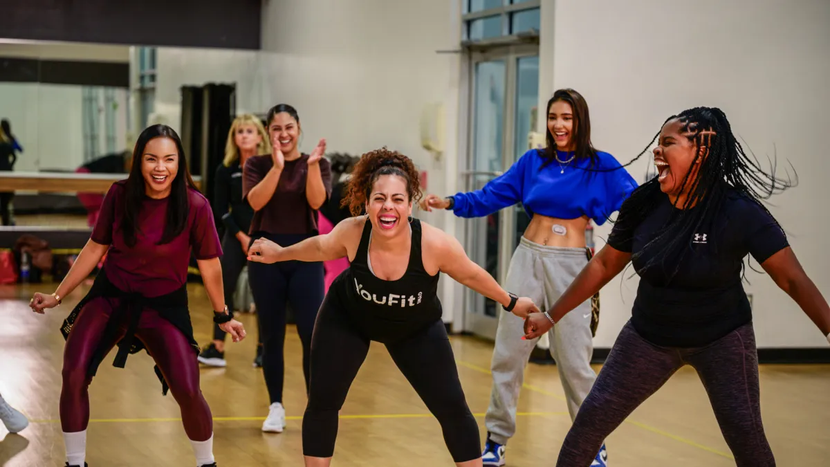 Diverse women enjoying a lively group fitness class, laughing and holding hands in a bright studio.