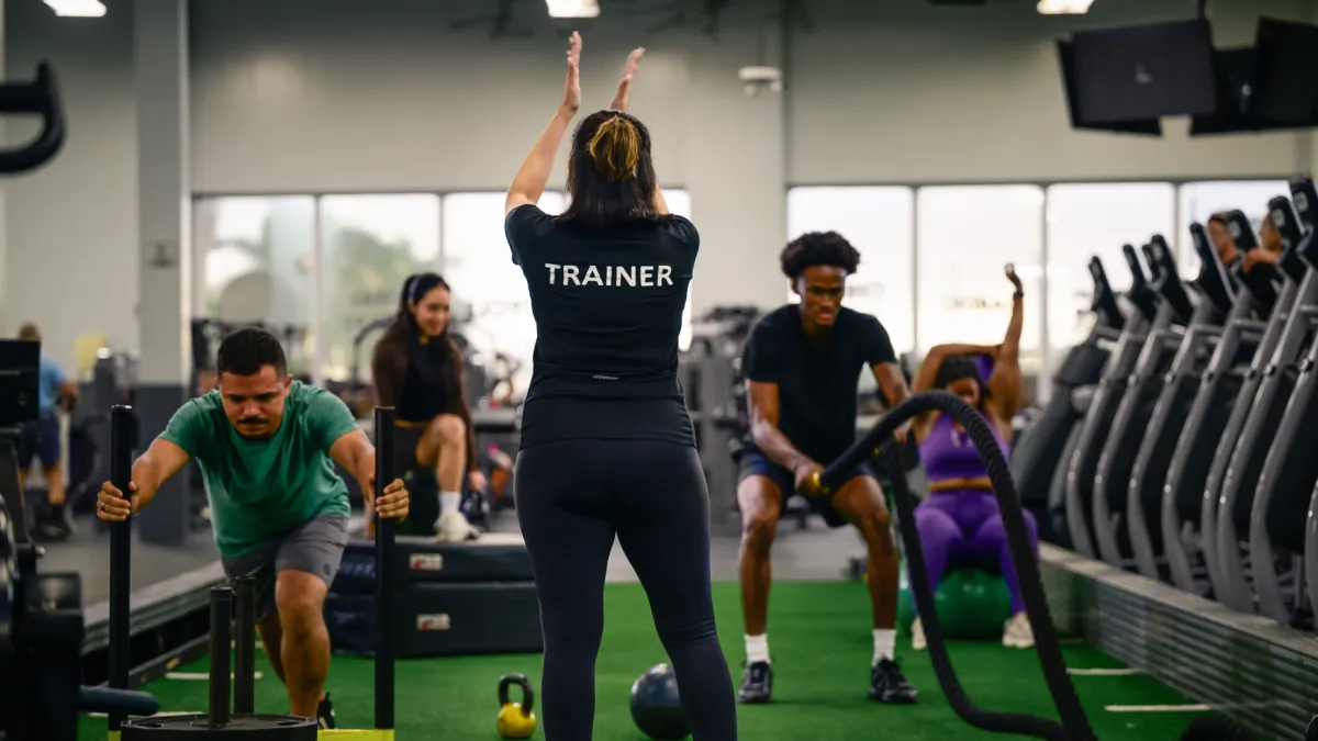 Fitness trainer leads group workout with sled push and battle ropes in a gym with green turf flooring.