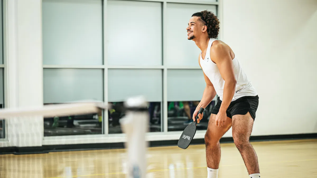Athletic man in white tank top and black shorts playing pickleball indoors with paddle in hand.