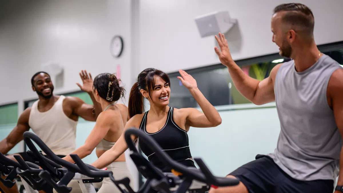 Group of diverse people smiling and high-fiving while cycling on stationary bikes in a gym class.