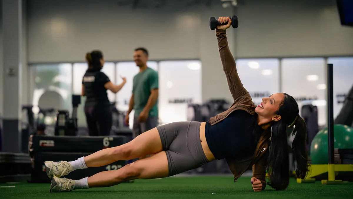 Young woman performs side plank with dumbbell in gym, two people chat in background near workout equipment.