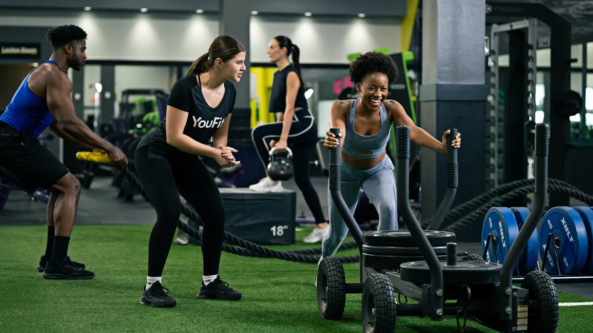 Diverse group training intensely in gym pushing weighted sled and performing kettlebell exercises with a trainer coaching.
